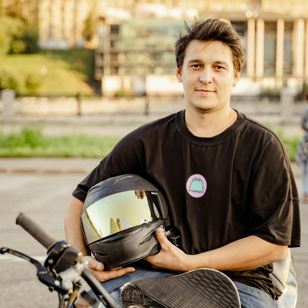 Man in black shirt holding helmet sitting on motorcycle in an outdoor setting.